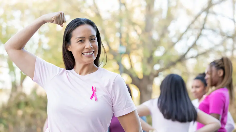 Woman wearing a pink ribbon flexes her arm in support of breast cancer awareness, with others in pink shirts in the background.