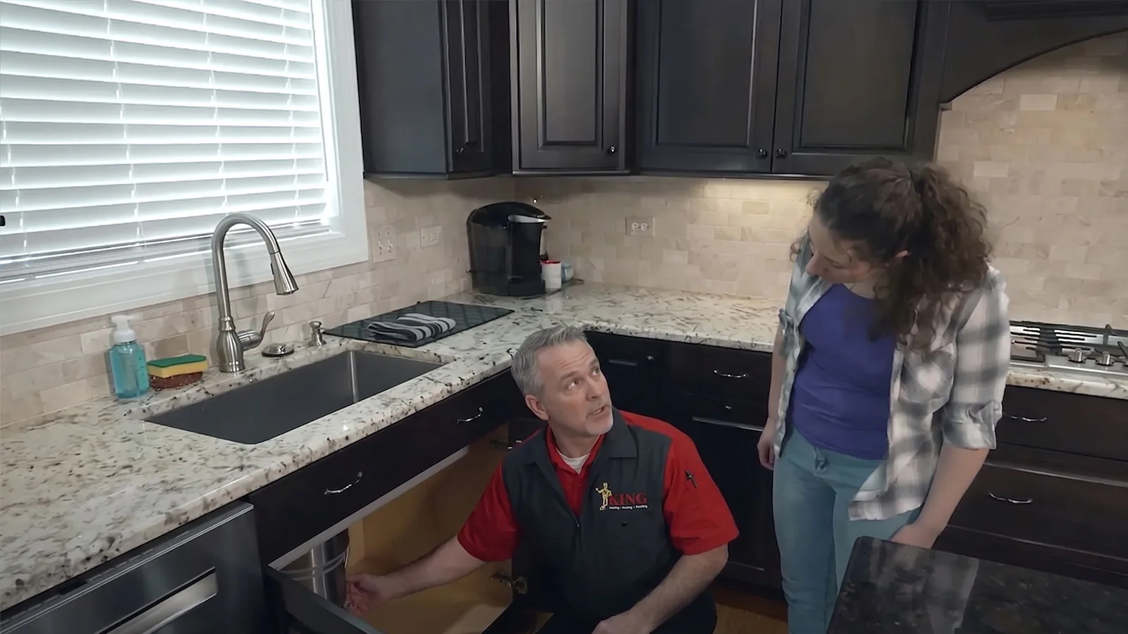 A King plumber working under a sink while a homeowner looks on